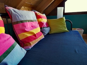 Een bed met een blauwe matras en kleurrijke gestreepte kussens in een kamer met houten balken en een blauwgroene wand.