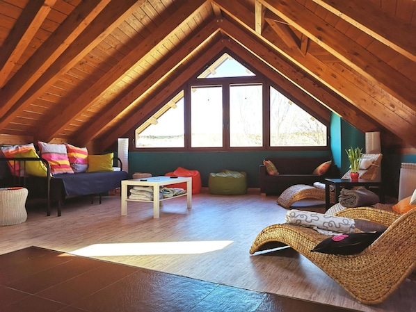 An attic room with a sloped ceiling, wooden beams, a large window, and a cozy seating area with cushions and a woven chair.