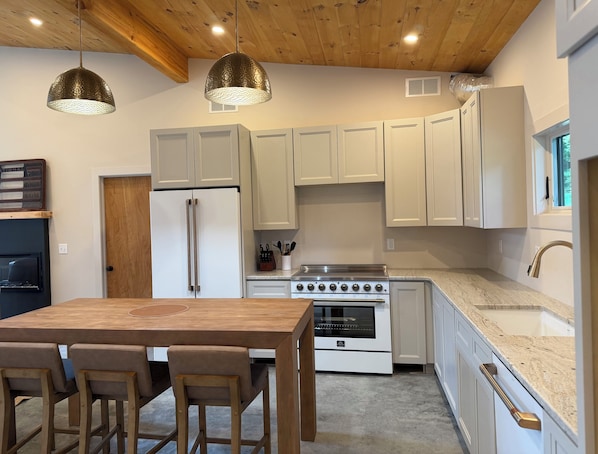 A modern kitchen with white cabinets, a stainless steel refrigerator, a gas stove, and a wooden dining table with bar stools.