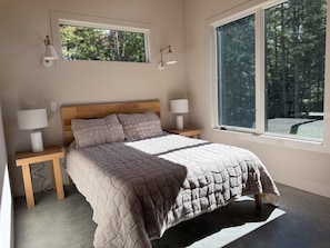 A bedroom with a wooden bed, two bedside tables with lamps, a large window with a view of trees, and a ceiling light fixture.
