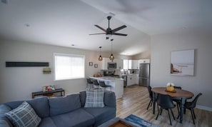 A modern living room with a gray sofa, a dining area with a wooden table and chairs, a kitchen with white cabinets, and a ceiling fan.