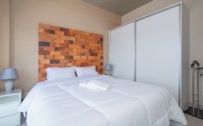 A hotel room with a bed, white linens, a brick-patterned headboard, a wardrobe, and a nightstand with a lamp.