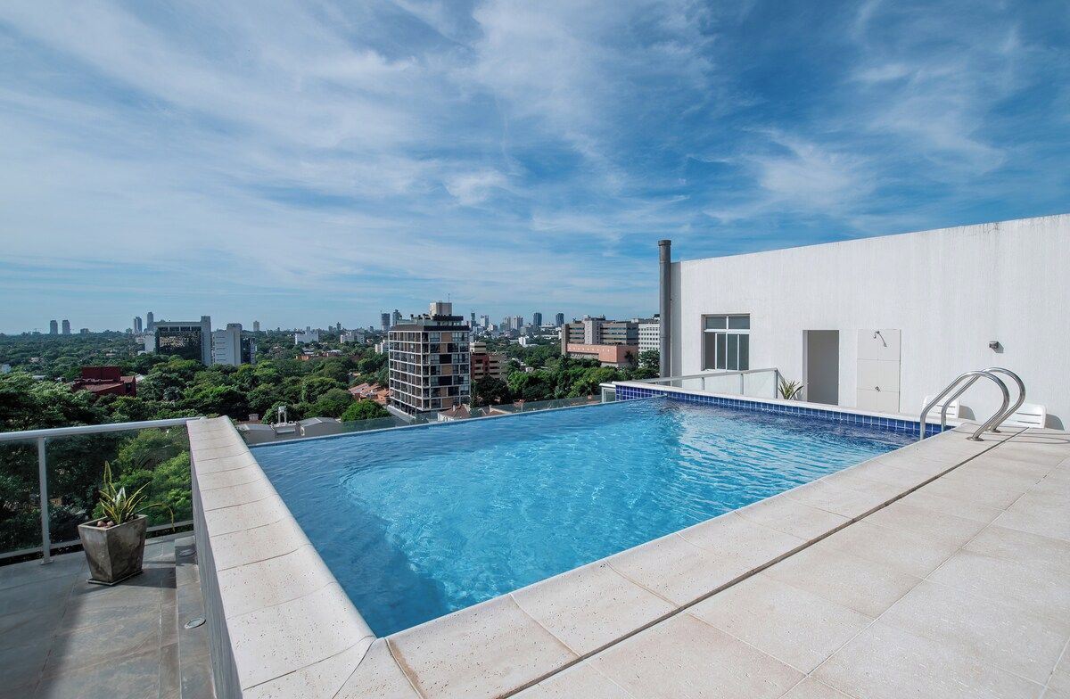 Piscina no terraço com vista para o horizonte urbano, um edifício branco e um guarda-corpo de metal.