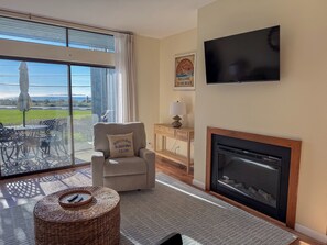 A living room with a fireplace, a television, a comfortable armchair, and a view of the outdoors. - Sunset Shores | Beach View Condo w/ Patio (Blaine)