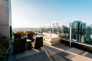 Terraza en la azotea con vistas a la ciudad, amueblada con sofás y sillones, y barandilla de cristal.