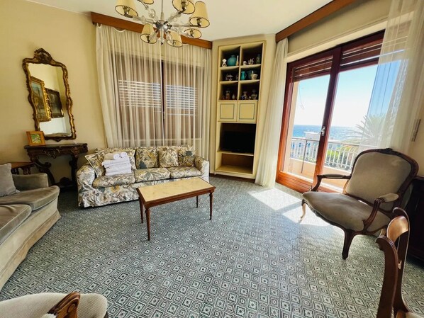 Una sala con un sofá estampado, una mesa de centro, una silla, un televisor y una estantería.