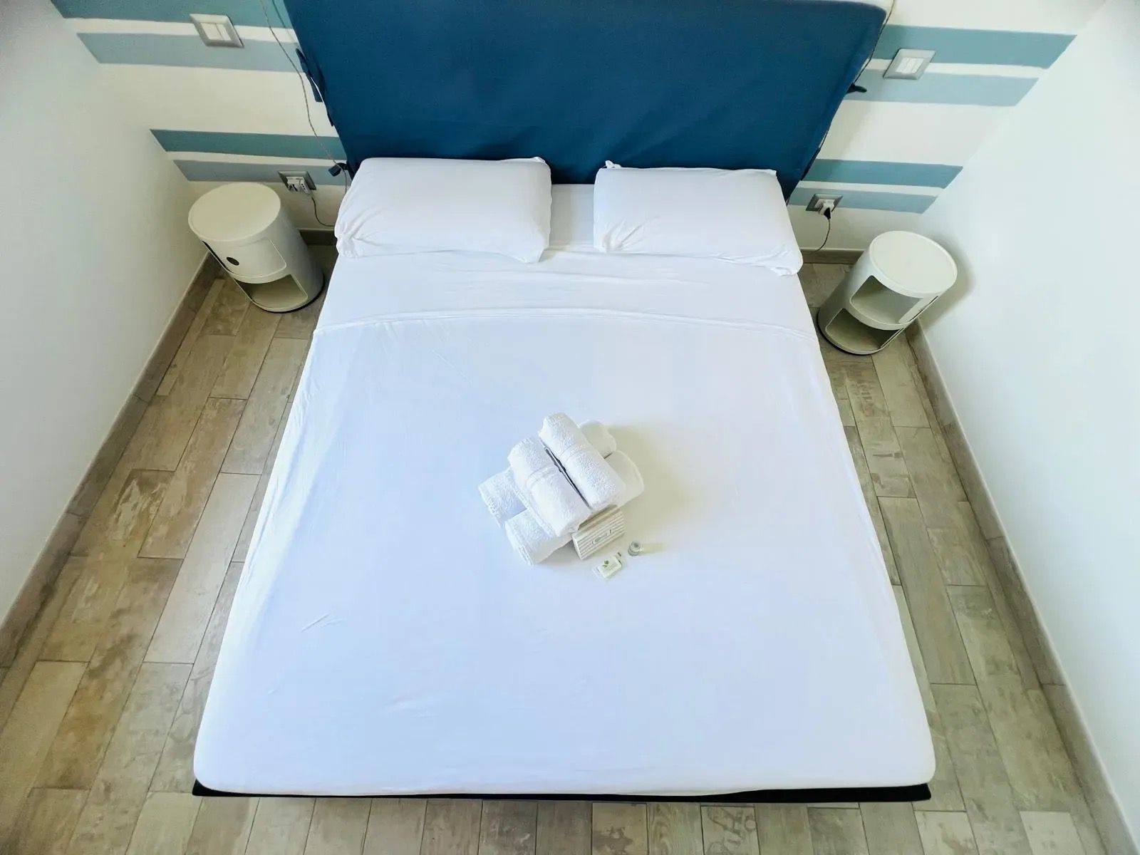 En pænt redt seng med hvide sengetæpper, en blå sengegavl og to natborde med lamper.