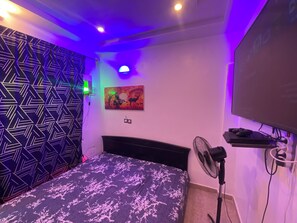 Quarto com cama, televisão na parede, um quadro e luminária no teto.