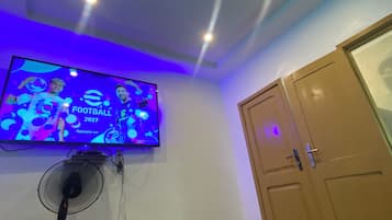 Huoneessa on sänky, seinälle asennettu televisio, jossa näkyy jalkapallo-ottelu, kattotuuletin ja ovi.