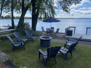 An outdoor seating area with Adirondack chairs, a fire pit, and a table with chairs, overlooking a lake.