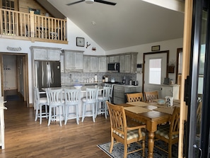 A spacious kitchen with a central island, wooden dining table, and chairs, with a view to the upper level.