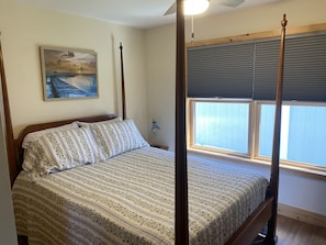 A four-poster bed with a floral-patterned bedspread, a painting on the wall, and a window with blinds.