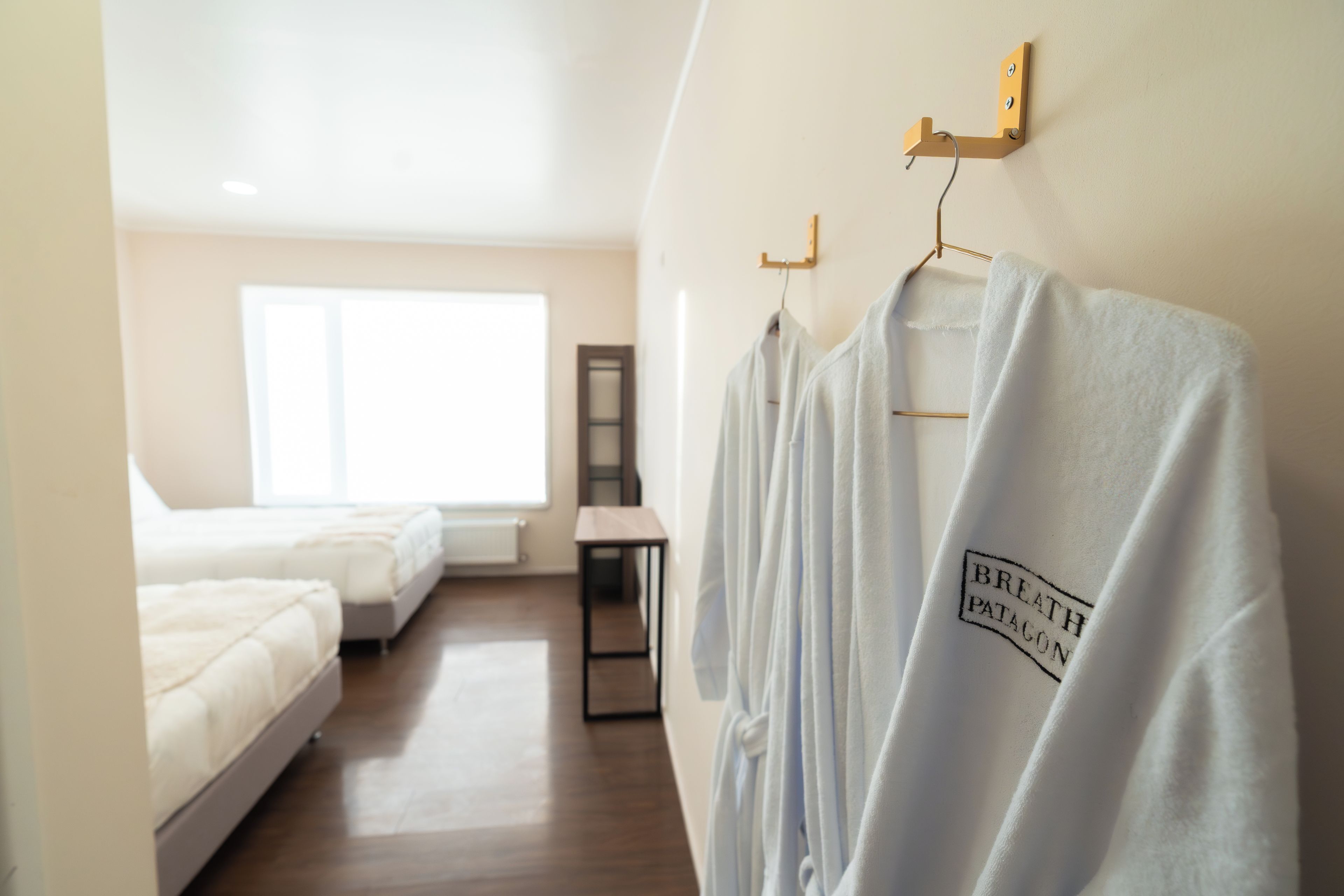 Habitación de hotel con dos camas, una mesita pequeña y un toallero de pared con toallas blancas.