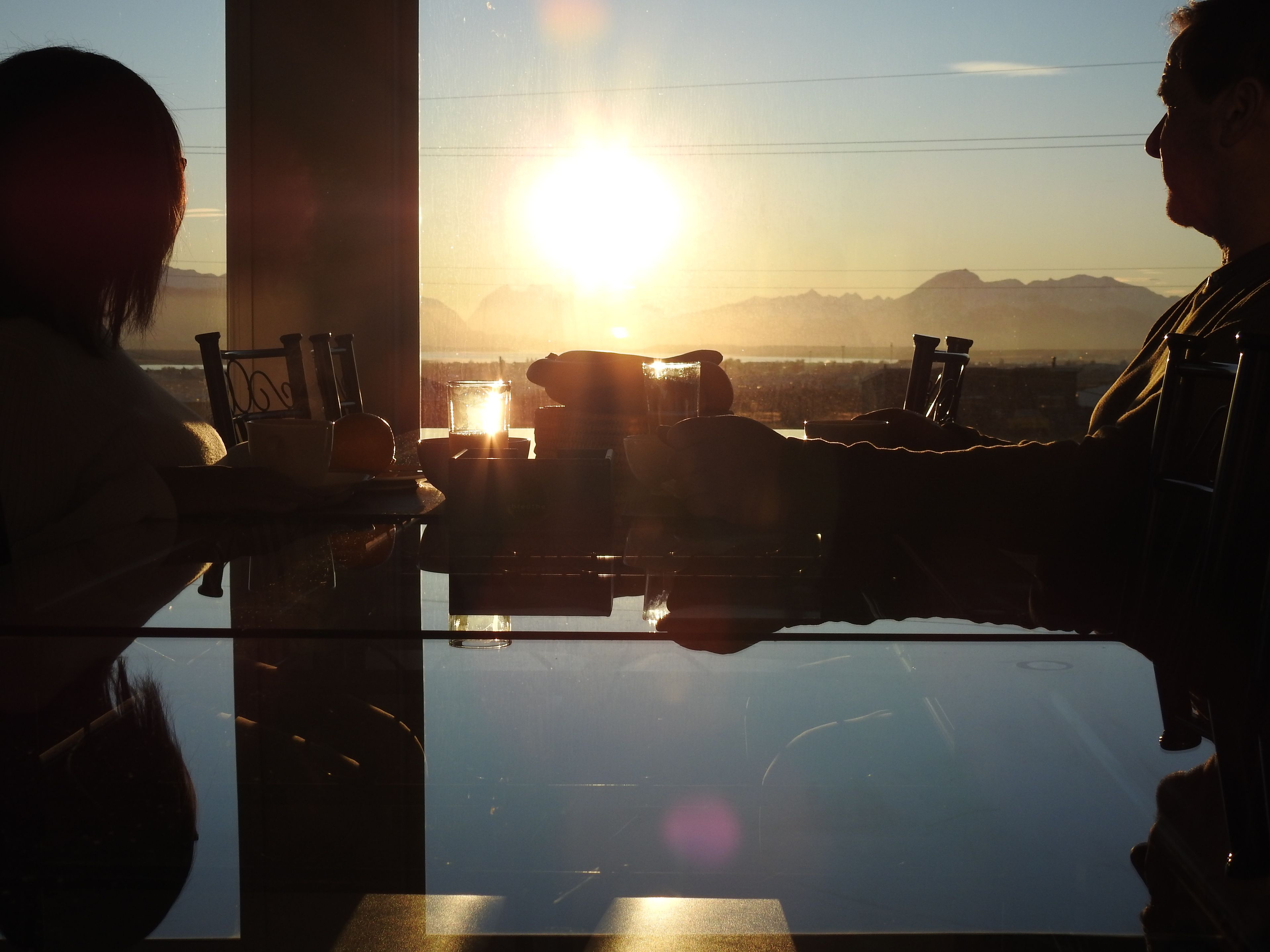 A dining area with a table and chairs, a glass partition, and a view of the sunset over mountains.