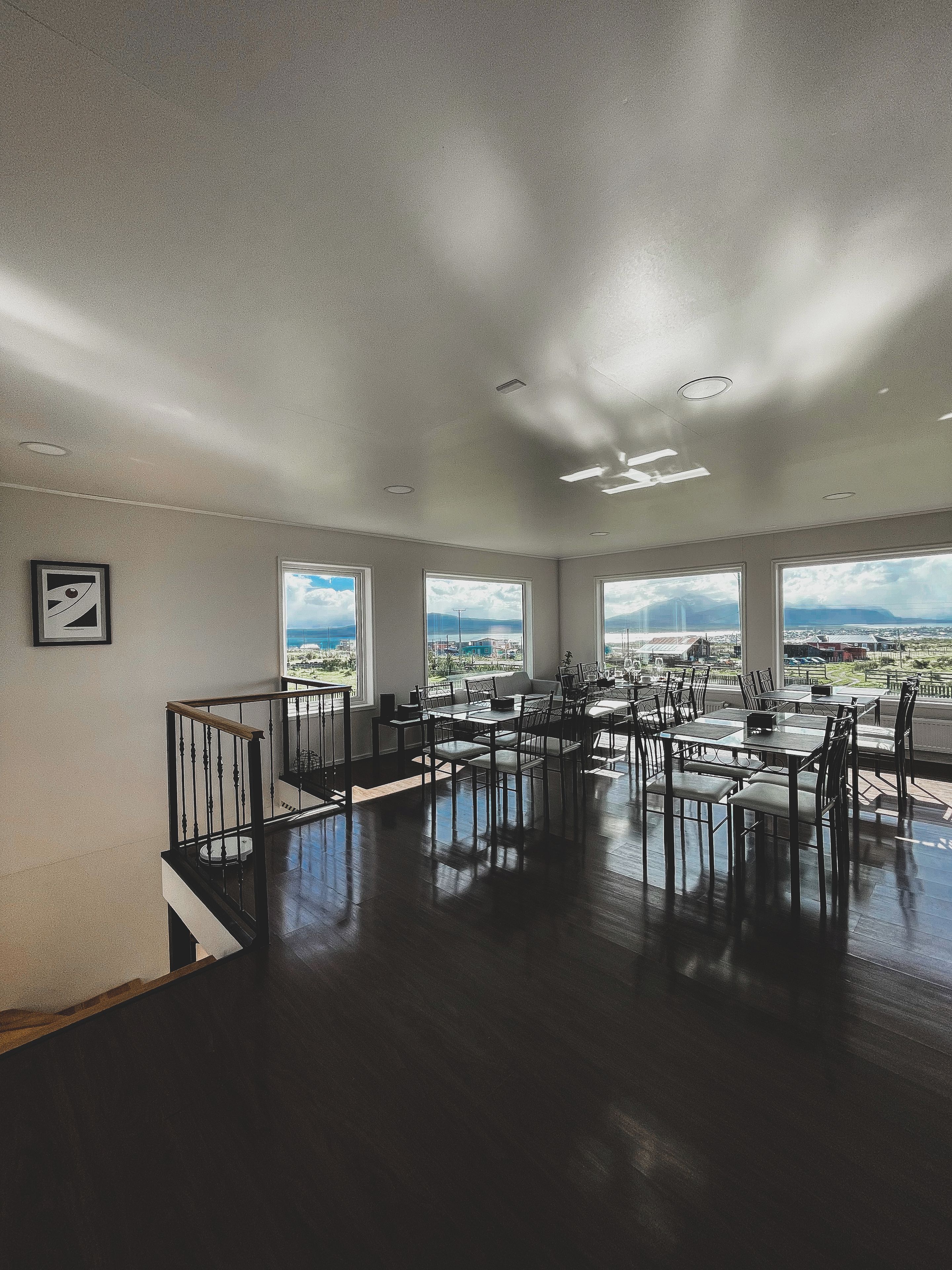 Un comedor amplio con varias mesas y sillas, ventanas grandes que ofrecen vistas del paisaje urbano y una escalera que conduce a un nivel superior.