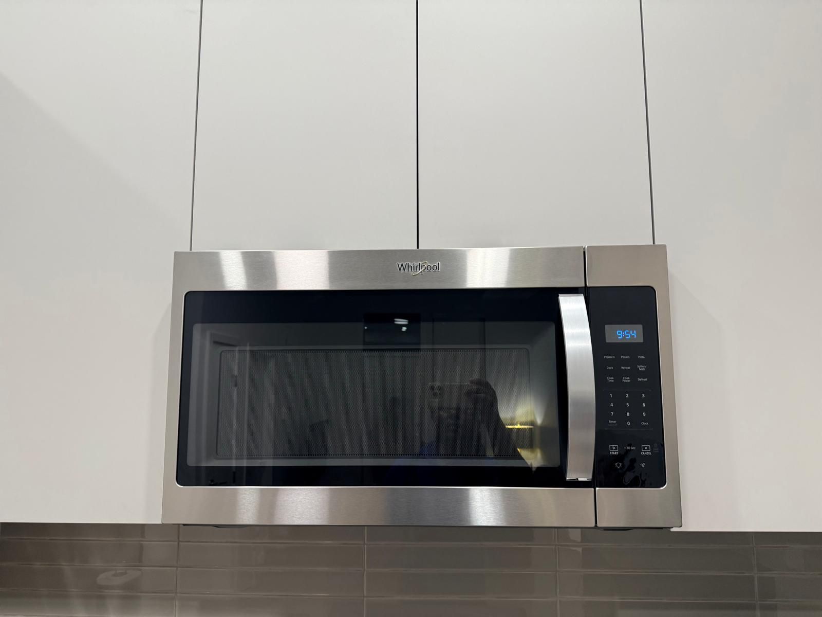 Eine Edelstahl-Mikrowelle von Whirlpool mit digitaler Anzeige, vor einem weißen Schrank und einer gefliesten Spritzschutzwand.