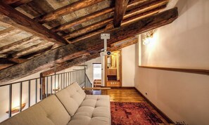Ein Loft-Zimmer mit freiliegenden Holzbalken, einer Couch, einem gemusterten Teppich und einer Treppe, die zu einer erhöhten Ebene führt.