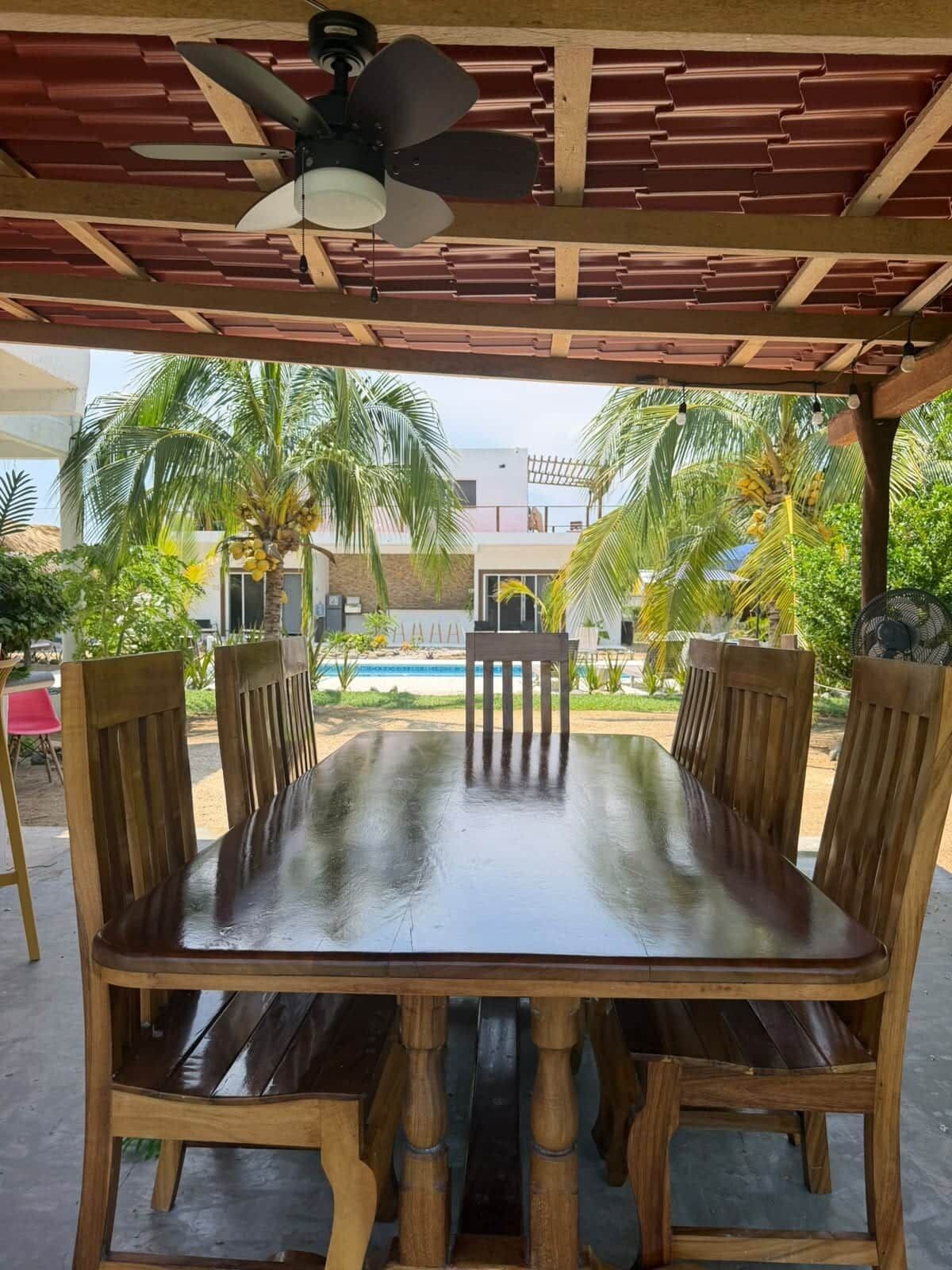 Ein Gartenmöbelset aus Holz mit Tisch und Stühlen unter einer Pergola mit Deckenventilator, umgeben von Palmen und einem Schwimmbad.