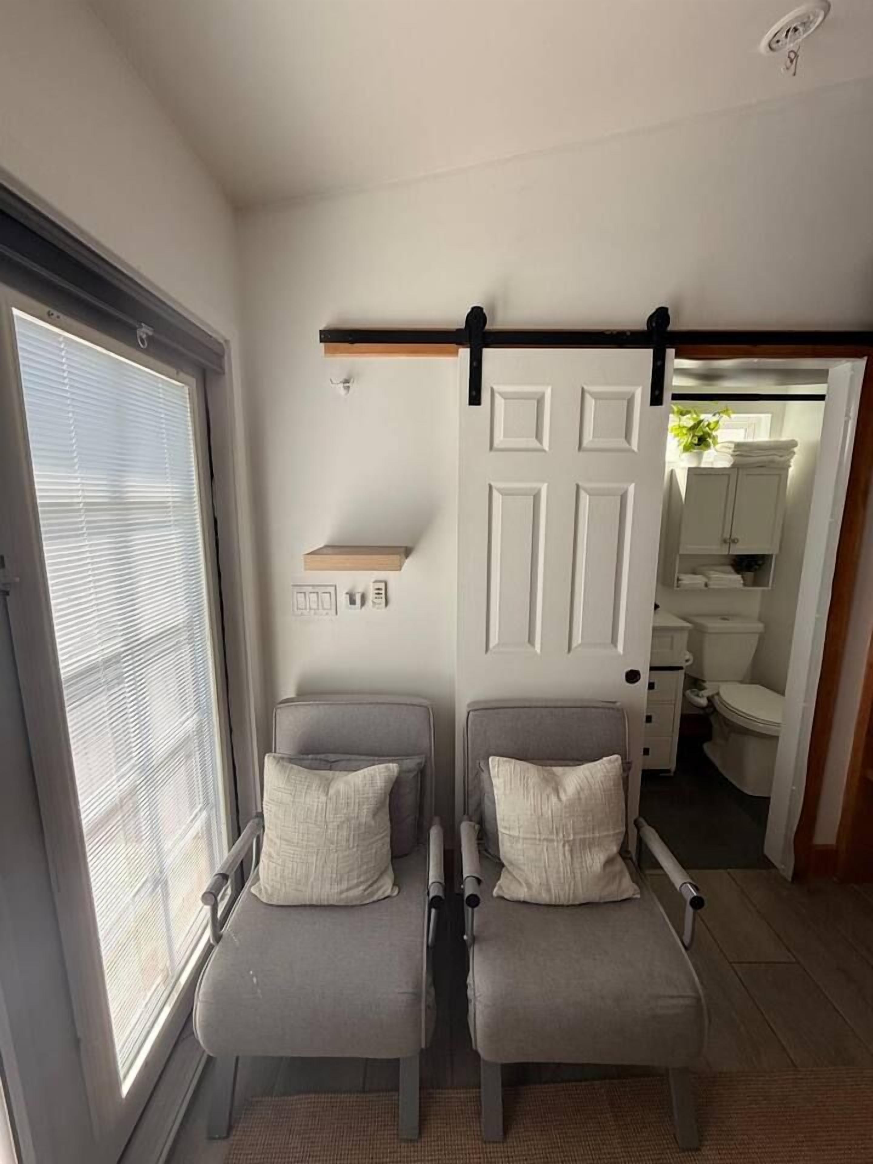 A room with two grey chairs, a white door, a window with blinds, and a bathroom visible through an open door.
