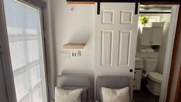 A room with two grey chairs, a white door, a window with blinds, and a bathroom visible through an open door.