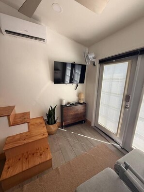 Una sala moderna con un televisor de pantalla plana montado en la pared, una escalera de madera, una pequeña planta y un cerramiento de vidrio corredizo que da a un espacio exterior.