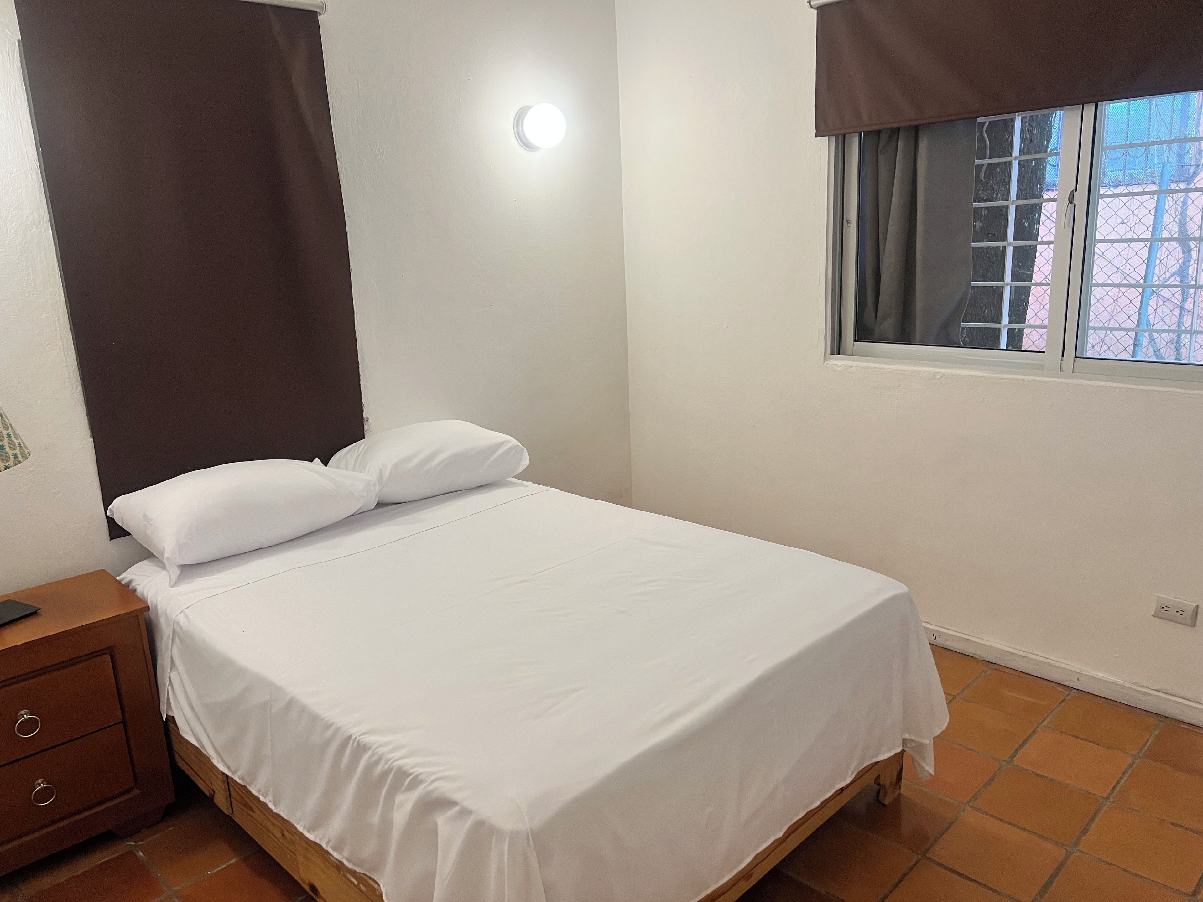 Una cama individual con ropa de cama blanca en una habitación con ventana, una mesita de noche y un enchufe.