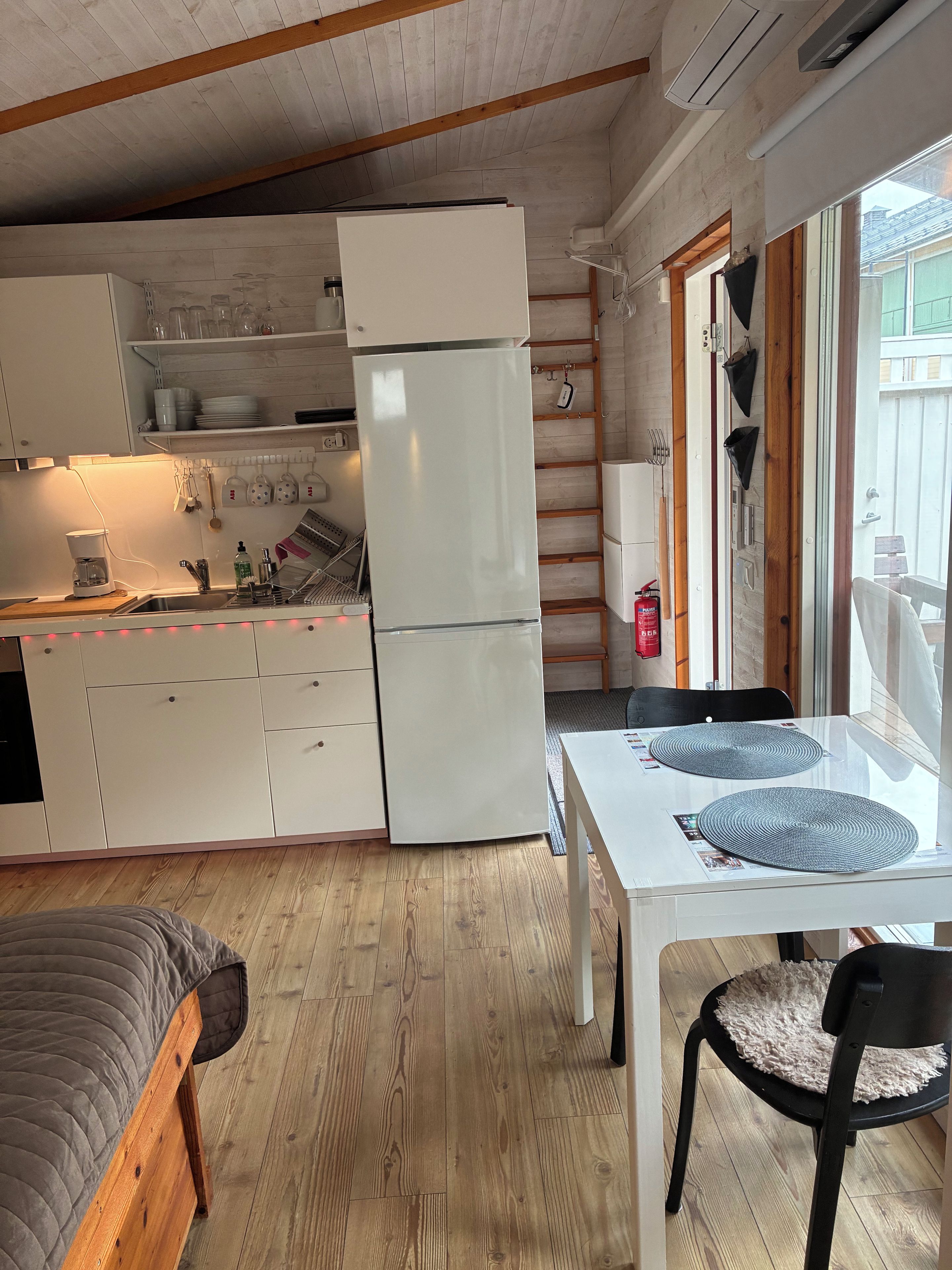 A compact kitchen with a refrigerator, sink, and wooden cabinets. There is a dining table with placemats and a chair.