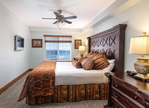Quarto de hotel com cama, cabeceira de madeira, ventoinha no teto, janela com persianas, luminária e televisão.