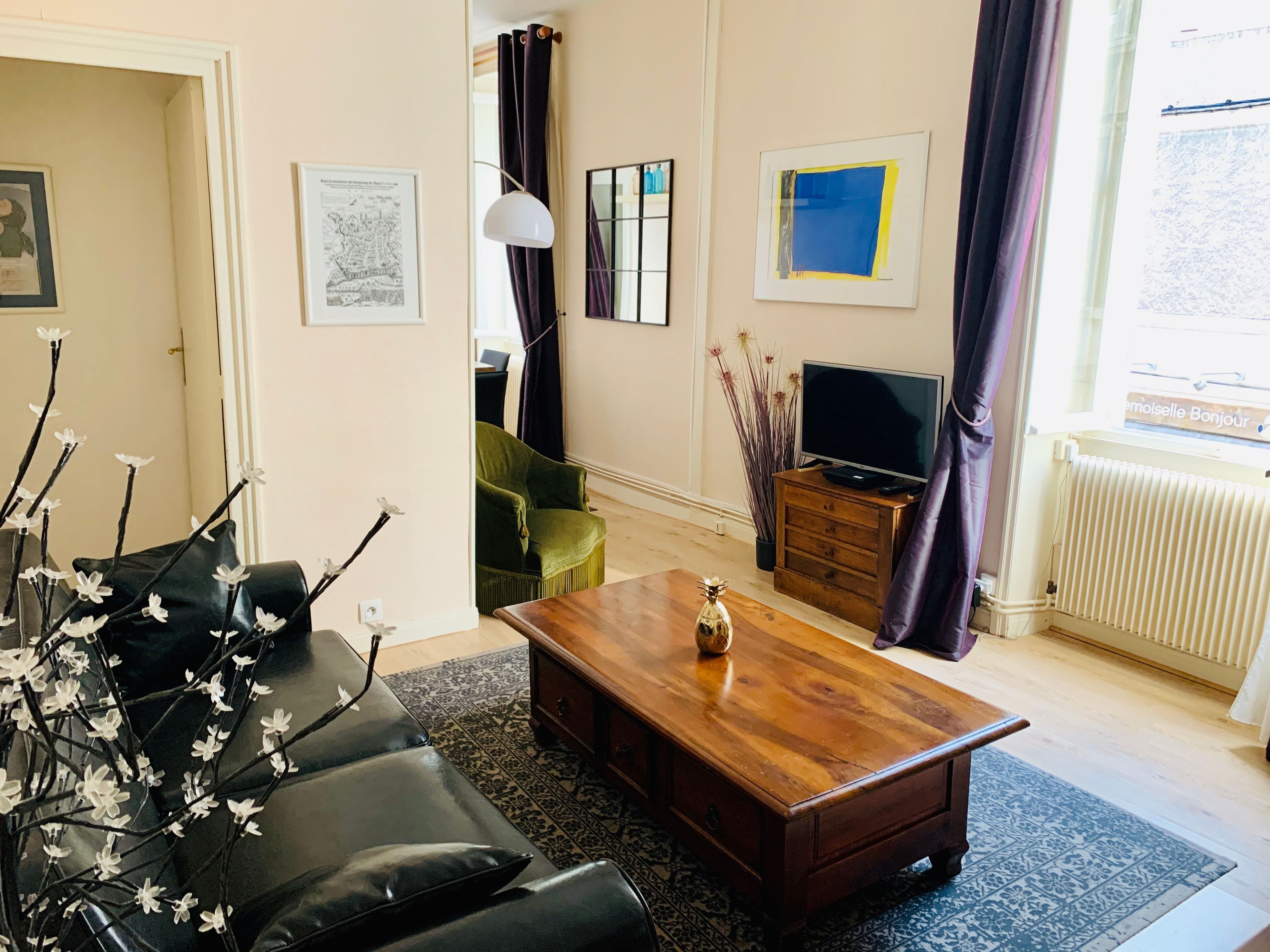 Ein Wohnzimmer mit einer Couch, einem Couchtisch und einem Fernseher. An den Wänden hängen Bilderrahmen, und ein Fenster ist mit Vorhängen versehen.