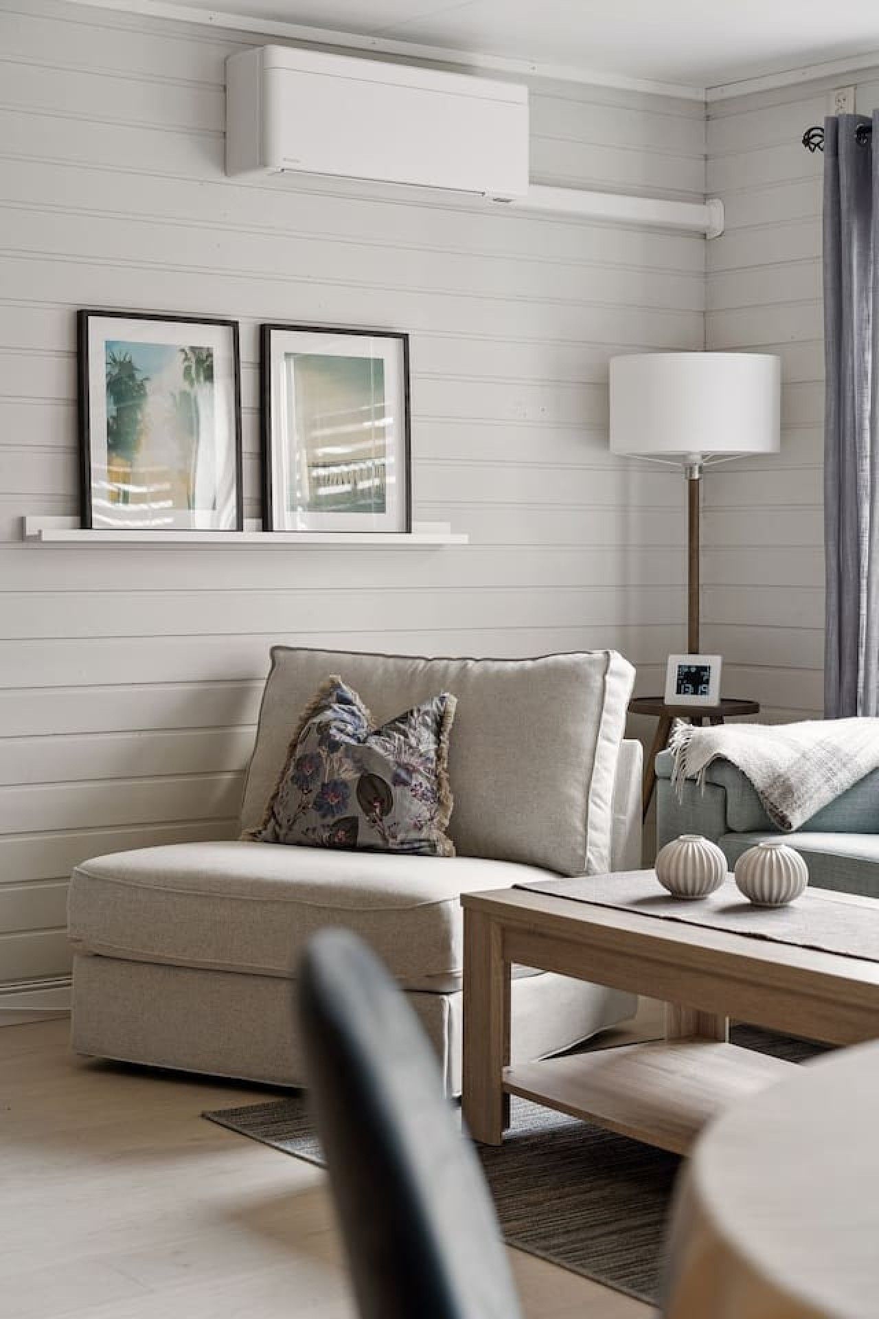 A modern living room with a sofa, a coffee table, and a floor lamp. There are two framed pictures on the wall and a small table with a clock.