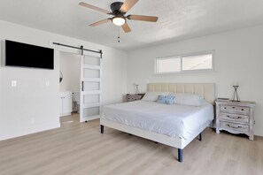 Een slaapkamer met een bed, nachtkastje, dressoir, plafondventilator, raam en deur.