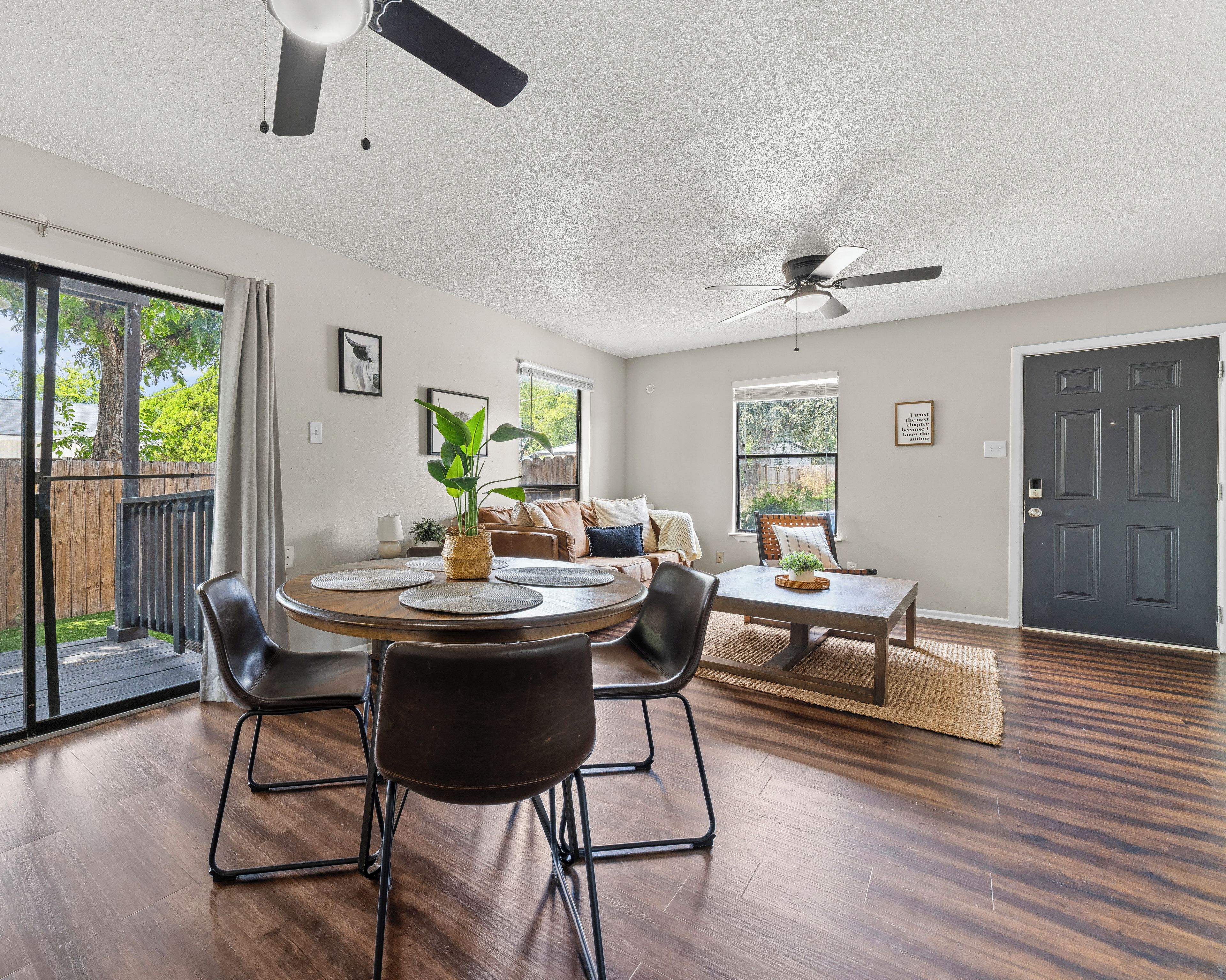 A spacious living room with a round dining table, black chairs, a ceiling fan, and a view of the outdoors through large windows.