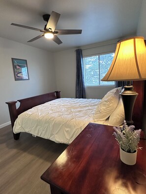 A bedroom with a bed, a wooden nightstand, a lamp, a ceiling fan, a window with curtains, and a wall art piece. - Modern 2-bedroom condo close to Hospitals and Shops-centrally located Rio Rancho (Rio Rancho)