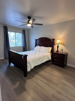 Ein Schlafzimmer mit einem Bett, einem Nachttisch, einer Lampe, einem Deckenventilator und einem Fenster mit Vorhängen.