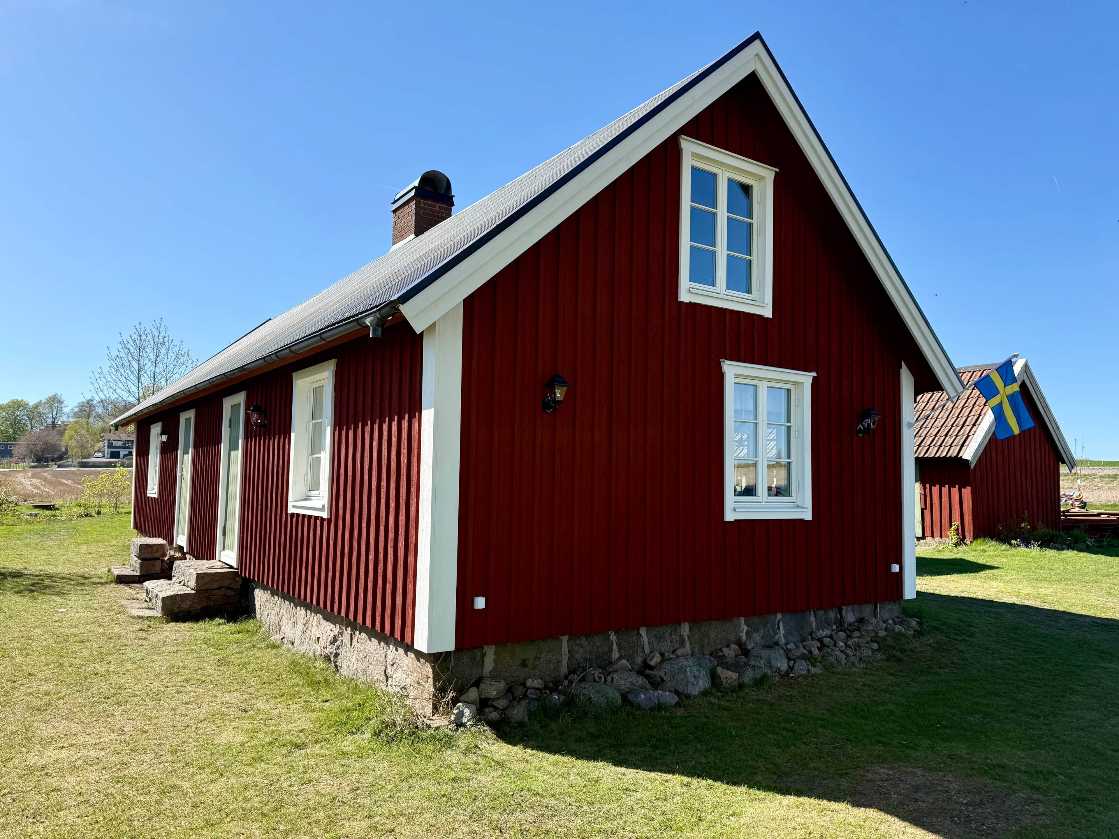 Une maison en bois rouge, avec des fondations en pierre, une cheminée et un drapeau.