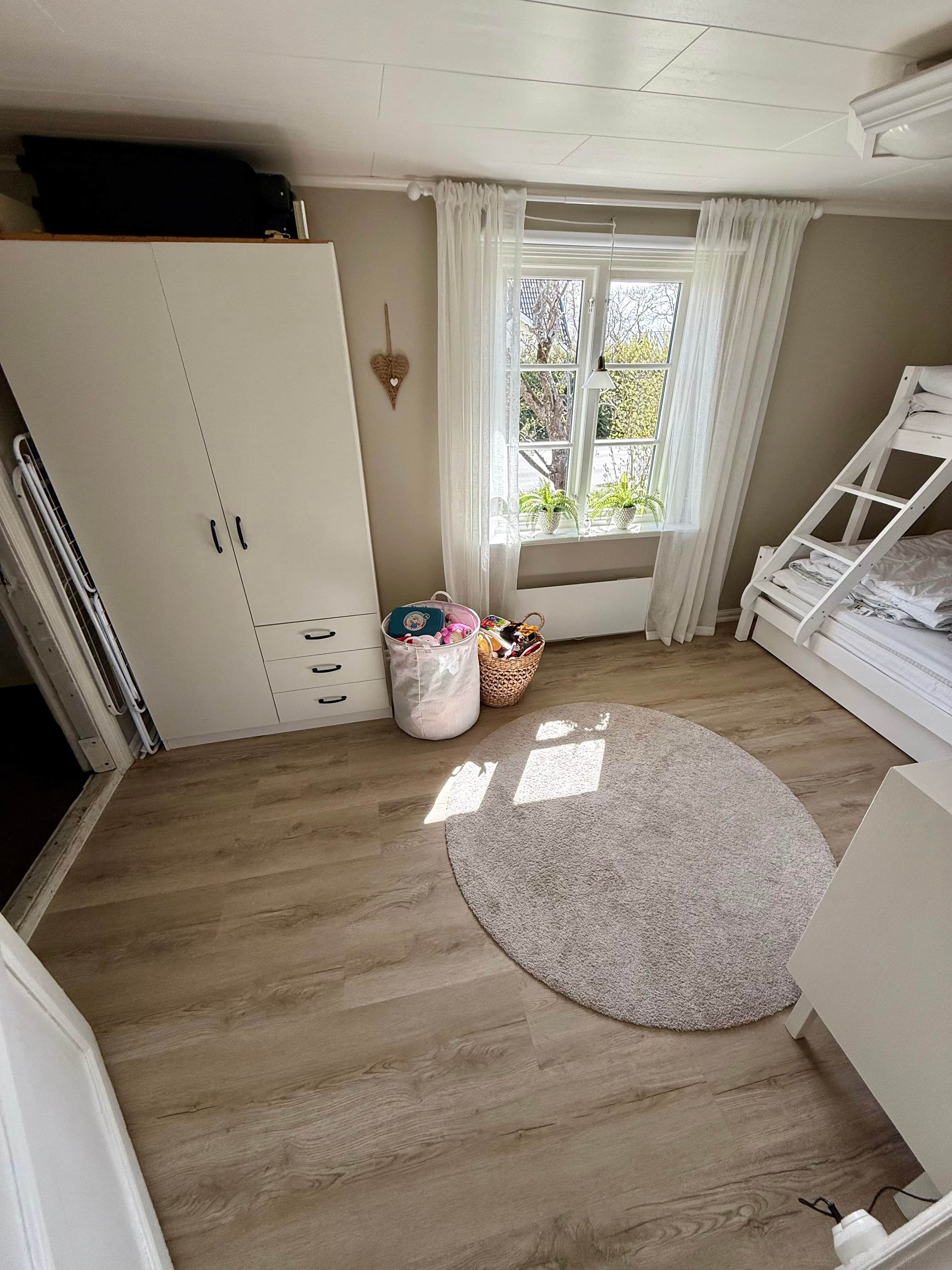 Une pièce avec un plancher en bois, une armoire blanche, une fenêtre avec des rideaux, un lit blanc et un tapis rond.