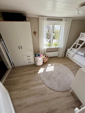 Una habitación con piso de madera, un armario blanco, una ventana con cortinas, una cama blanca y un tapete redondo.