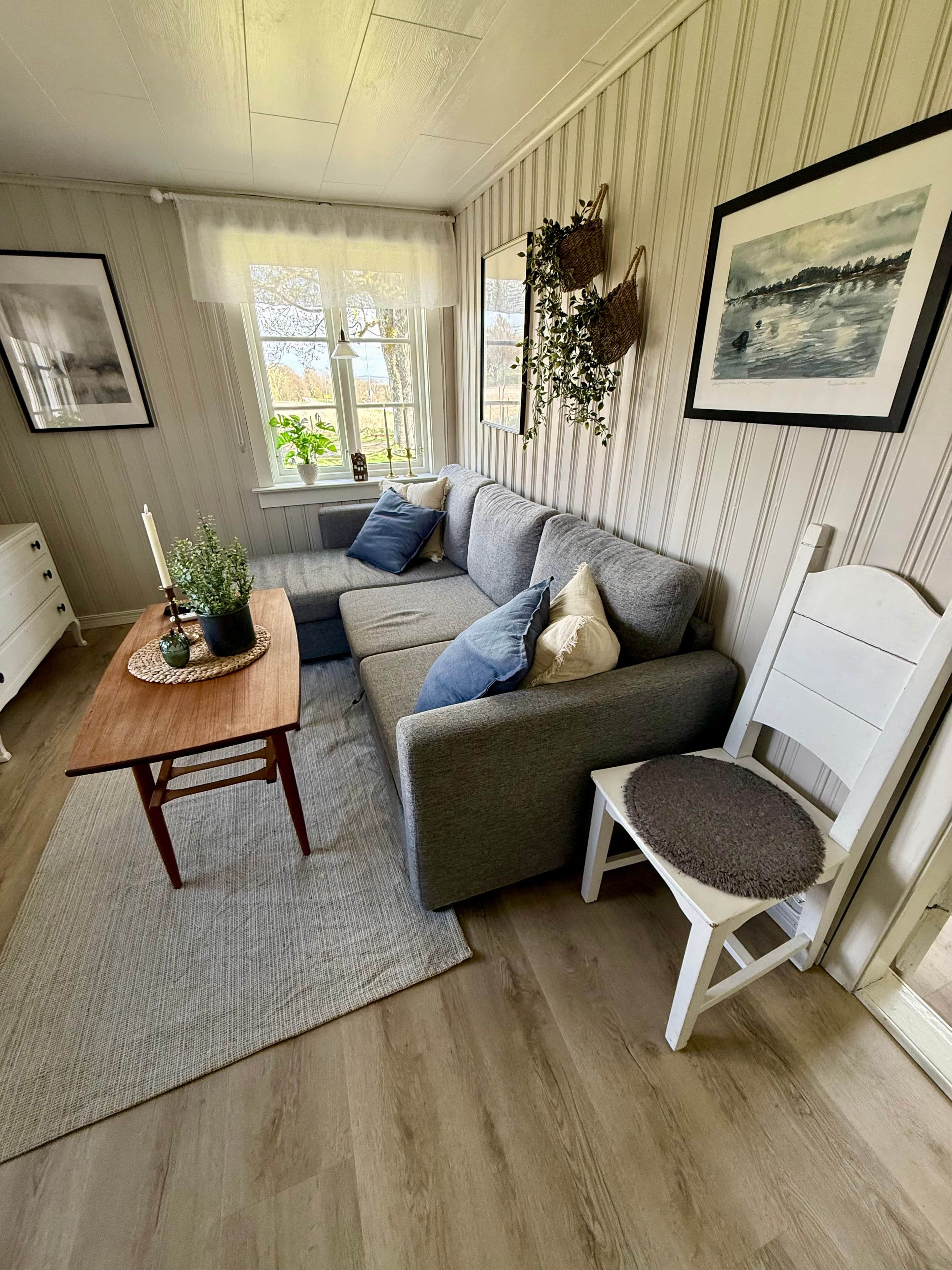 A cozy living room with a grey sofa, a wooden coffee table, and a white chair. There is a window with a view of greenery outside.