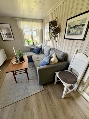 Una sala de estar acogedora con un sofá gris, una mesa de centro de madera y una silla blanca. Se ve vegetación a través de una ventana.