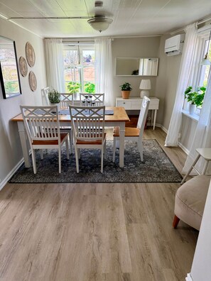 Un comedor con mesa de madera, sillas blancas, alfombra negra y planta en maceta.