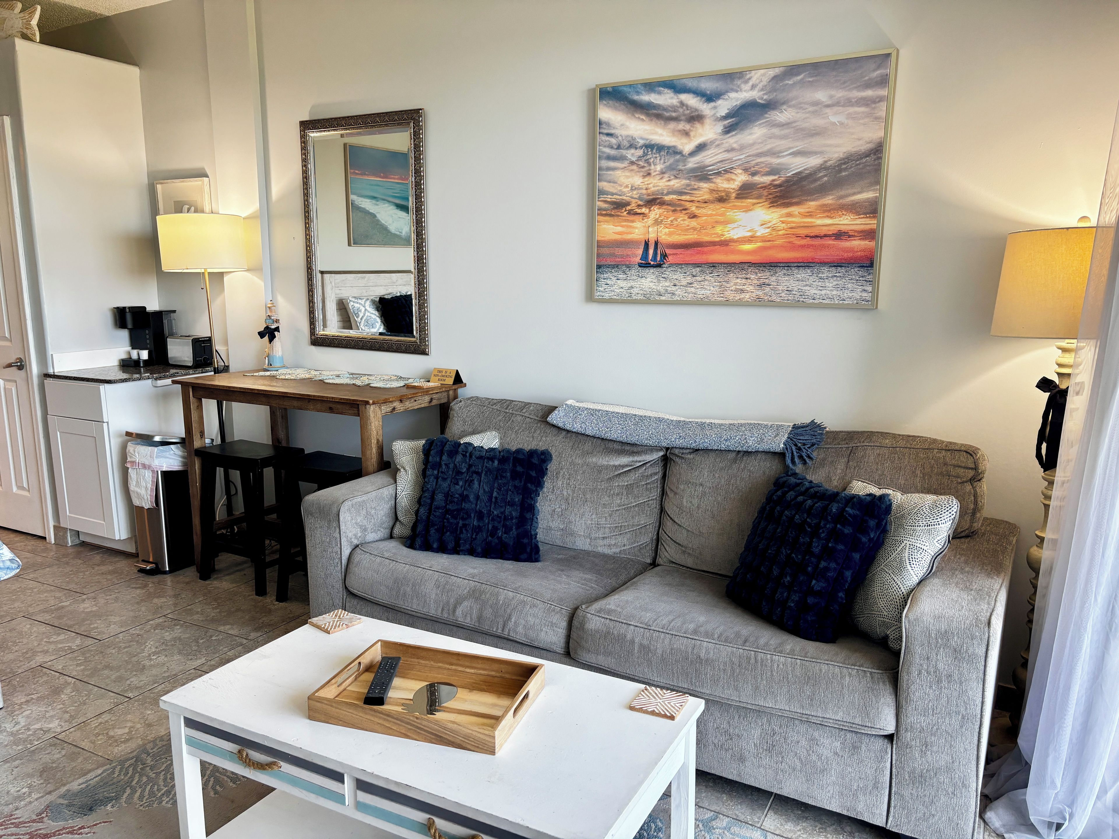 A living room with a grey sofa, a coffee table, a dining table with chairs, a kitchen area, and a painting of a sunset.