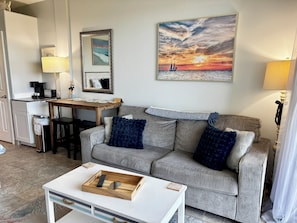 Ein Wohnzimmer mit grauem Sofa, einem Couchtisch, einem Esstisch mit Stühlen, einer Küchenzeile und einem Sonnenuntergangsbild.