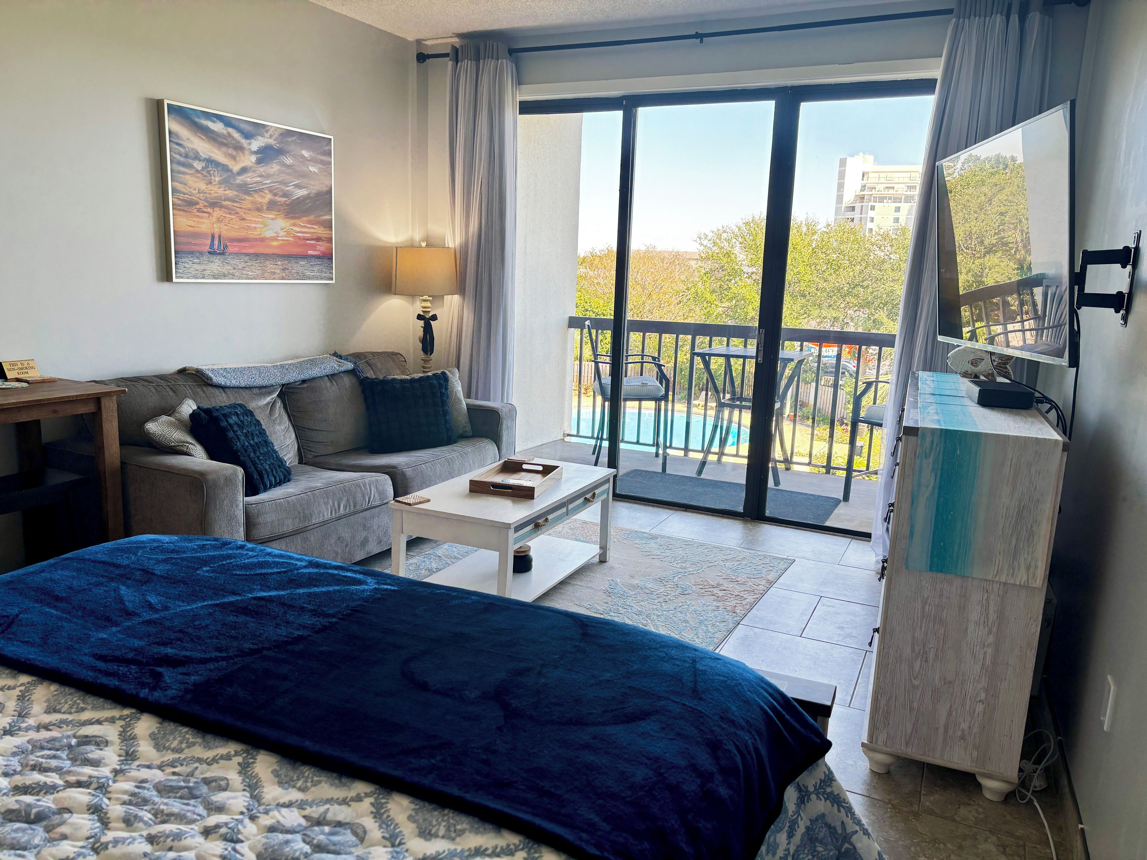 A hotel room with a sofa, a coffee table, a bed, a TV, and a balcony with a pool.
