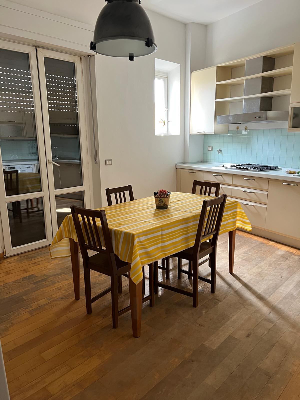 A dining area with a table set for four, wooden chairs, a black pendant light, and a kitchen with white cabinets and a tiled backsplash.