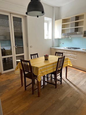 Un'area pranzo con tavolo apparecchiato per quattro persone, sedie in legno, una lampada a sospensione nera e una cucina con pensili bianchi e zoccolo piastrellato.