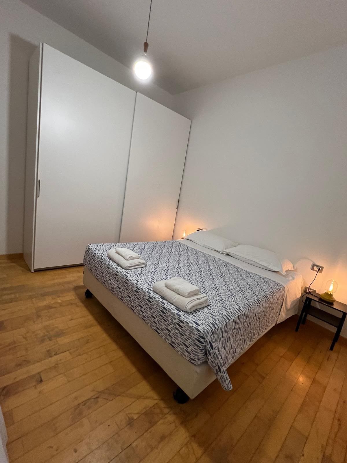 A bedroom with a bed, a wardrobe, a bedside table with a lamp, and a wooden floor.