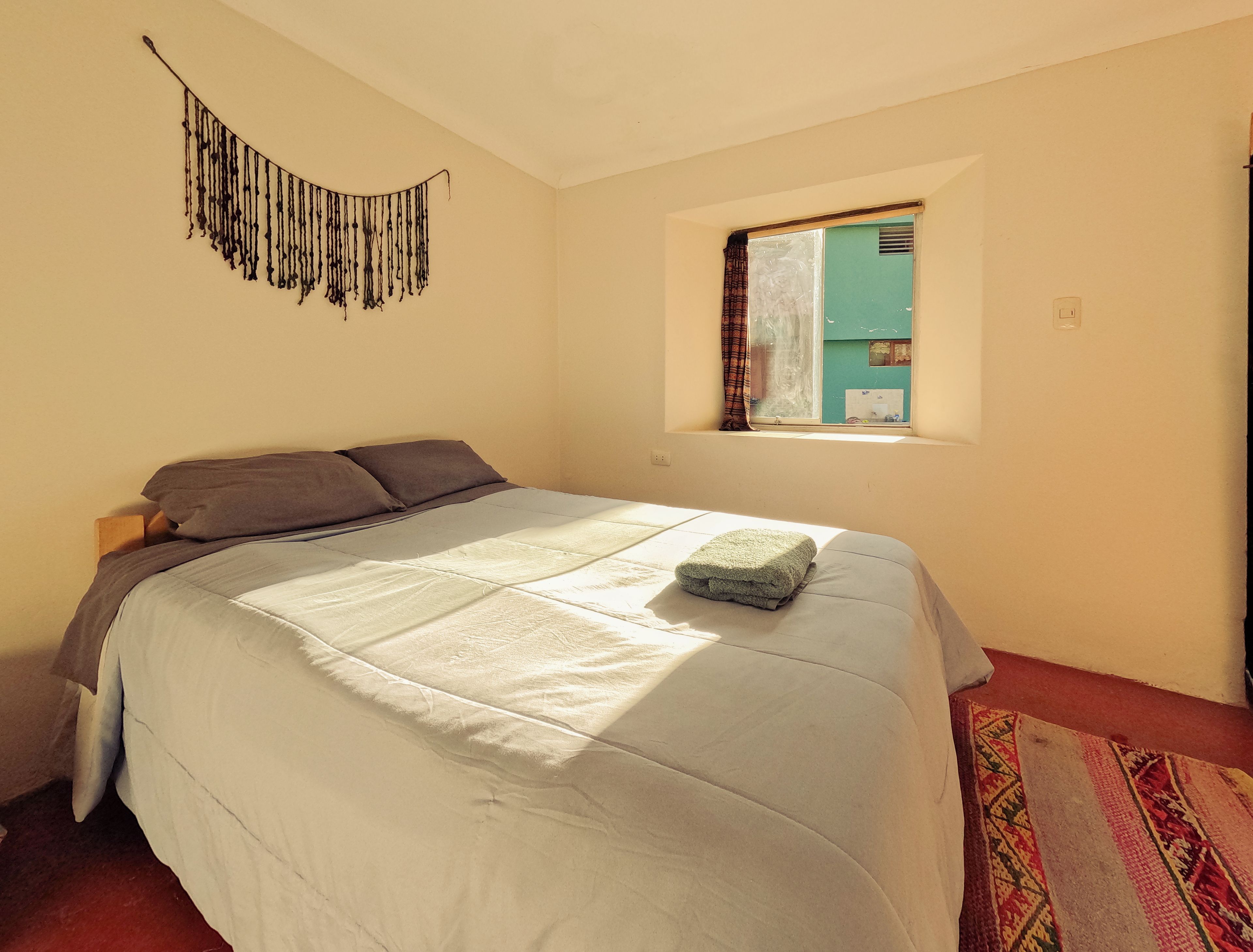 A bedroom with a bed, a window with curtains, a wall hanging, and a patterned rug.