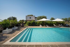 Piscina com espreguiçadeiras e guarda-sóis, rodeada por vegetação e um edifício.