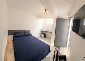 Et soveværelse med seng, lille skrivebord, stol, ventilator og garderobeskab.
