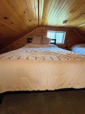 A bedroom with a sloped ceiling, a bed with white bedding and rose petals, a window with a view of trees, and a nightstand with a lamp.